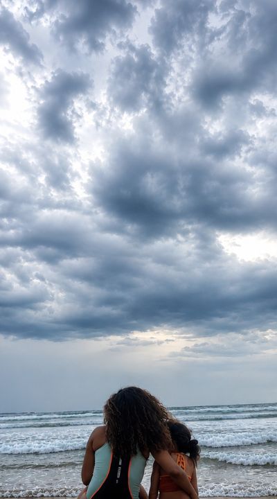 Two people sitting together on the shore looking out at the ocean under a cloudy sky
